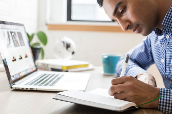 Student writing in a notebook at a desk with a laptop computer open